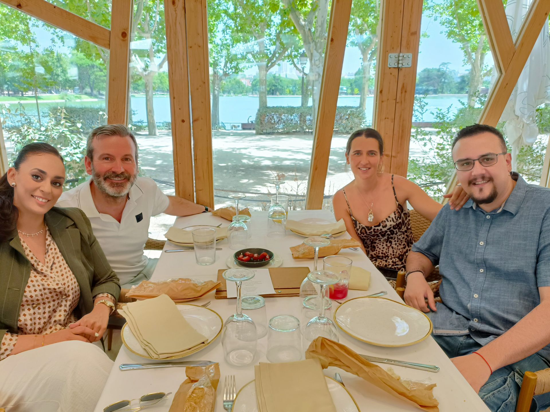 Cuatro personas sentadas a una mesa, comiendo al aire libre bajo una estructura de madera con vistas al agua.