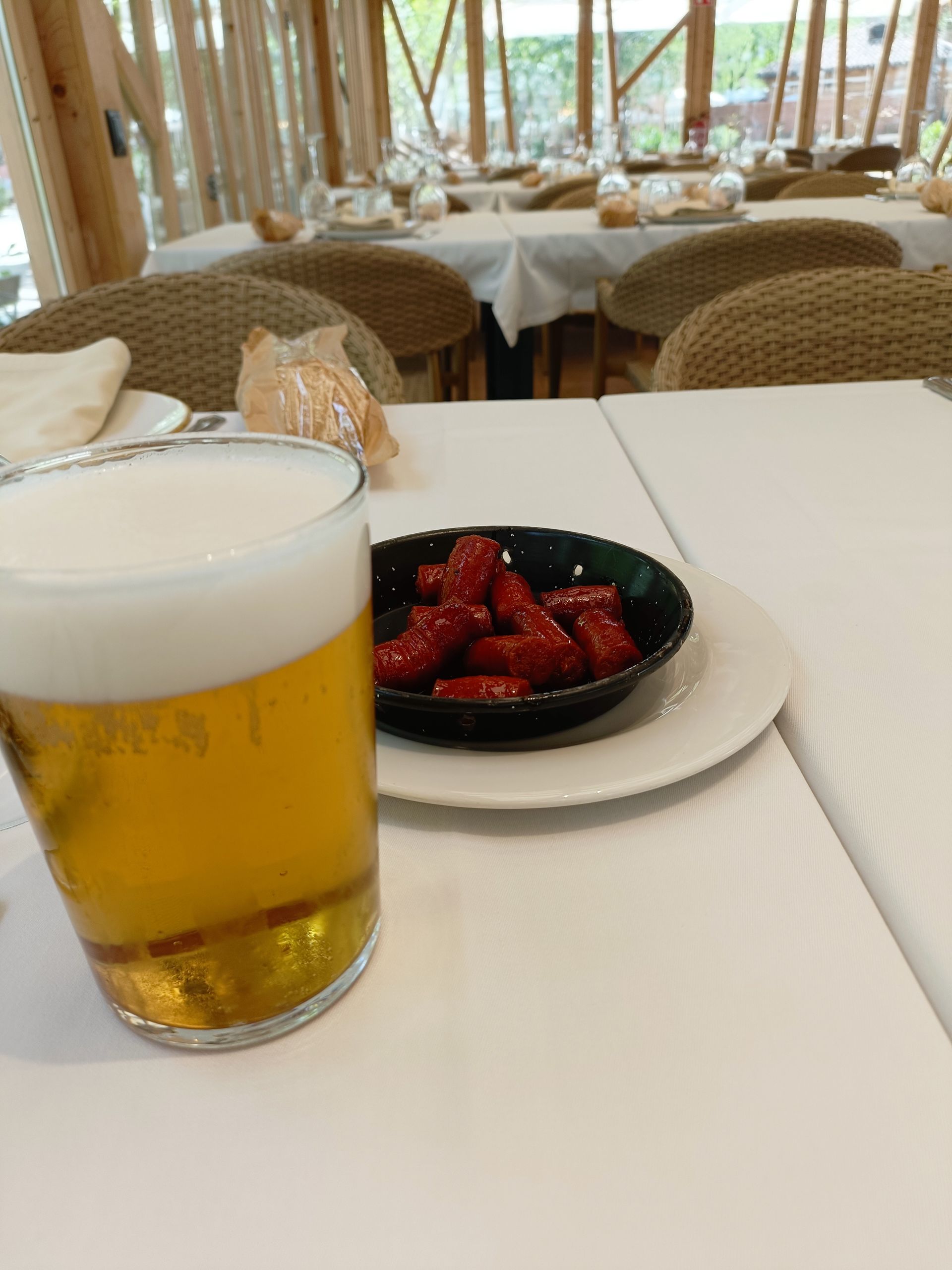Un vaso de cerveza y un bol de aperitivos rojos en un restaurante con mesas blancas.