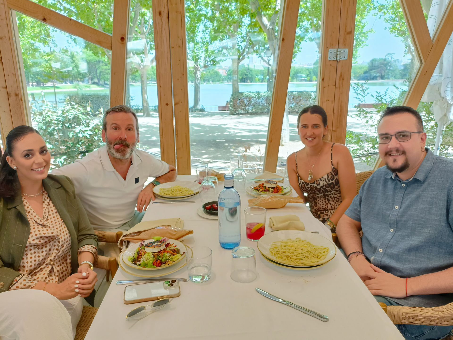 Cuatro personas están sentadas a una mesa en un luminoso restaurante al aire libre con vistas al agua, disfrutando de una comida.