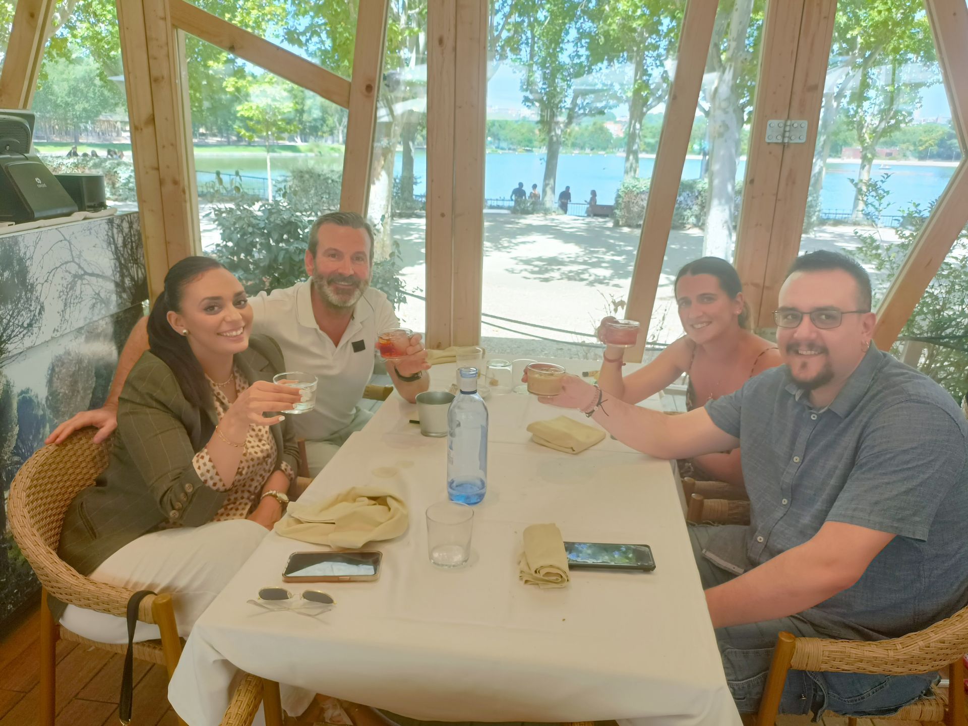 Cuatro personas brindando con bebidas en una mesa junto a una ventana con vistas a un lago.