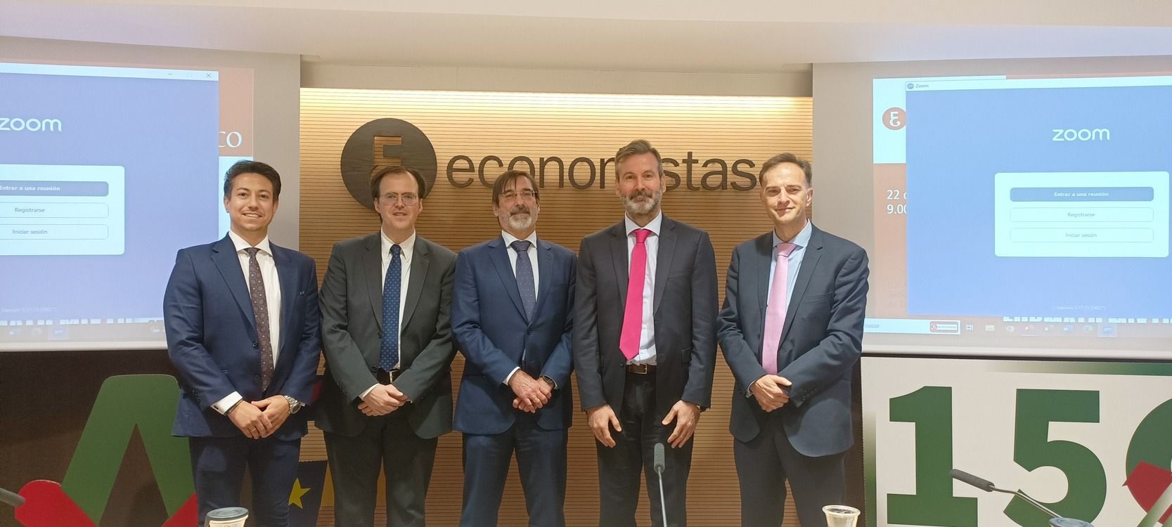 Cinco hombres de traje están de pie frente a un fondo con un logotipo. Detrás de ellos hay una pantalla con la palabra Zoom.