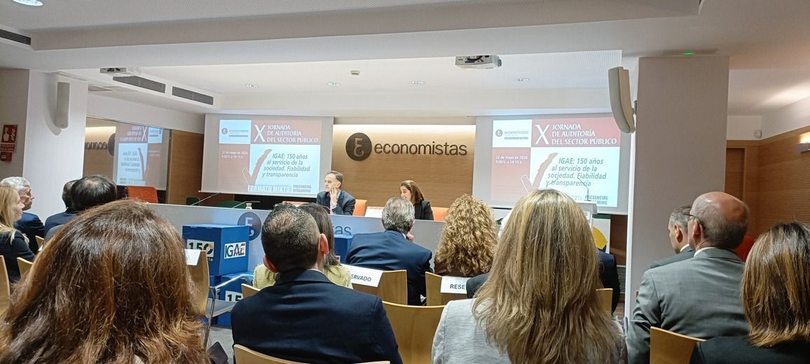 Público sentado en una conferencia, diapositivas de la presentación en la pantalla. Un orador está de pie al frente.