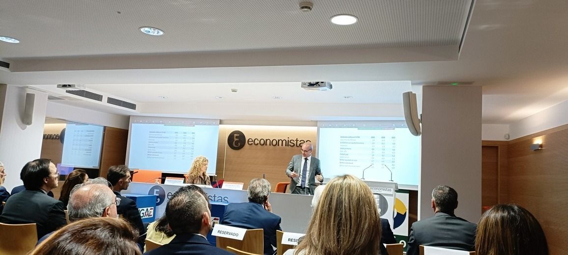 Una presentación en una sala de conferencias. Un orador se encuentra de pie frente a una pantalla con un gráfico; un público sentado escucha.