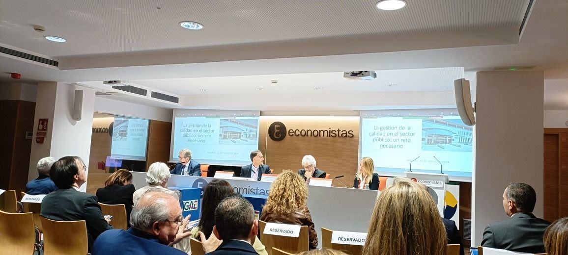 Una conferencia o reunión en una sala grande, con oradores en un escenario, público sentado y presentaciones en pantallas.