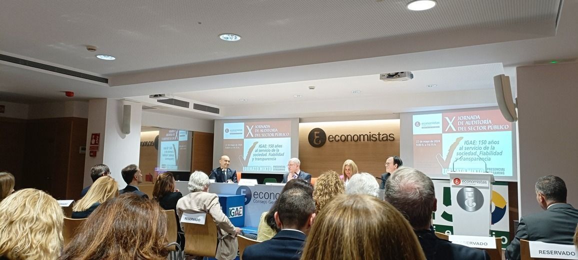 En una conferencia, el público escucha a los oradores desde un podio. Las pantallas de presentación son visibles.