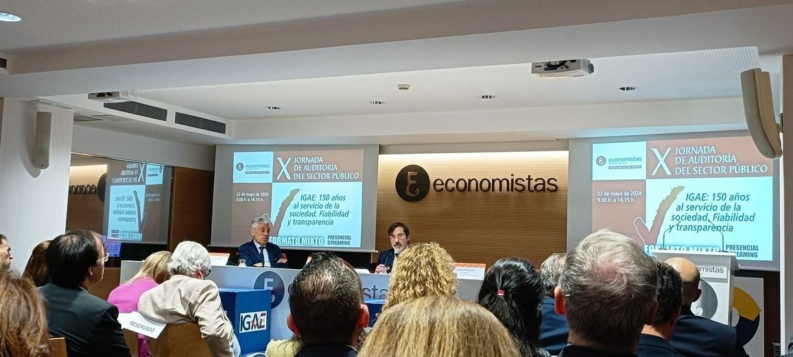 Una presentación en un auditorio: los ponentes sentados a una mesa, el público en filas, dos grandes pantallas detrás de ellos.