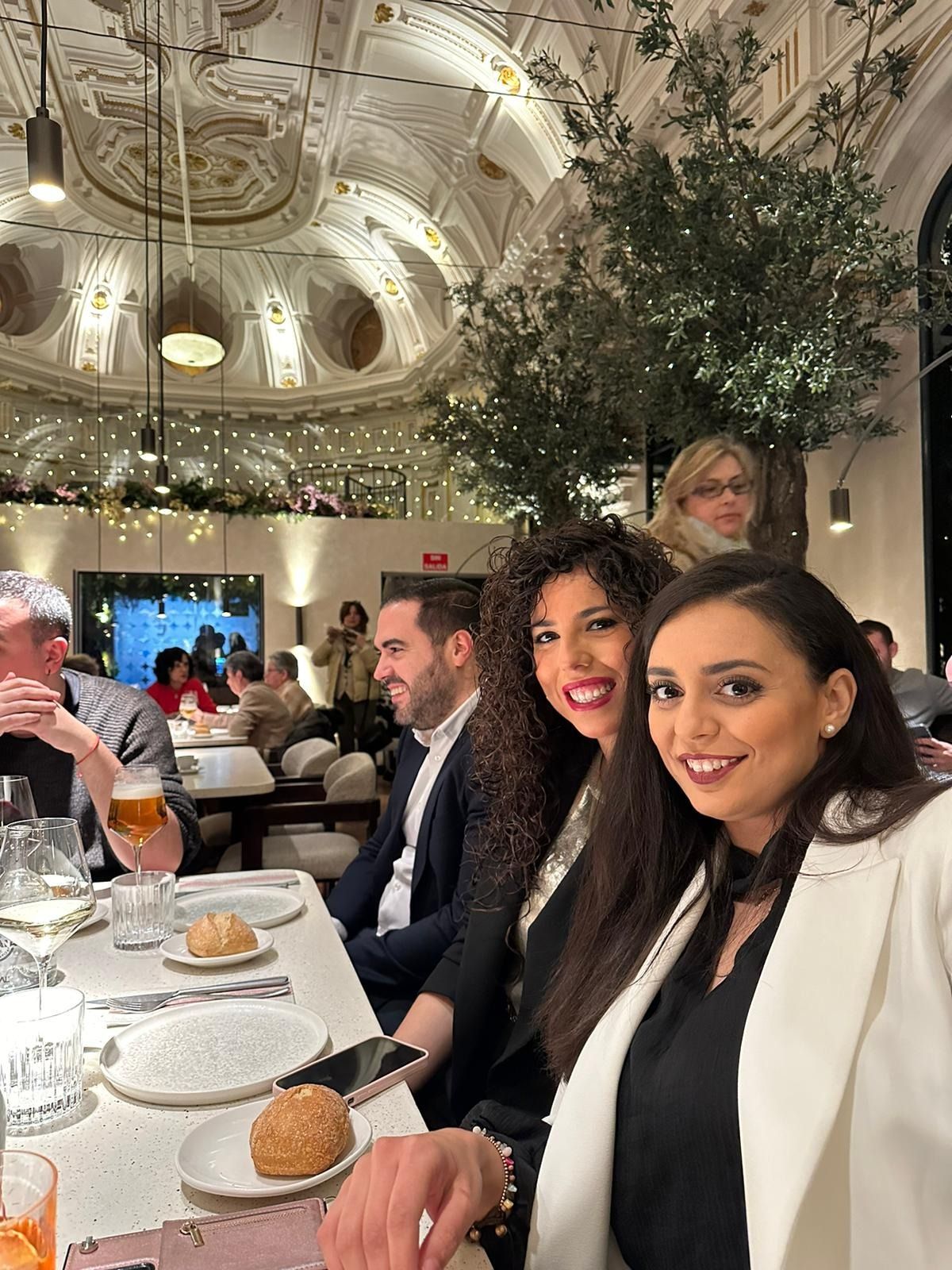 Dos mujeres sonríen en una mesa de un restaurante. Otras personas cenan al fondo. Techo ornamentado.