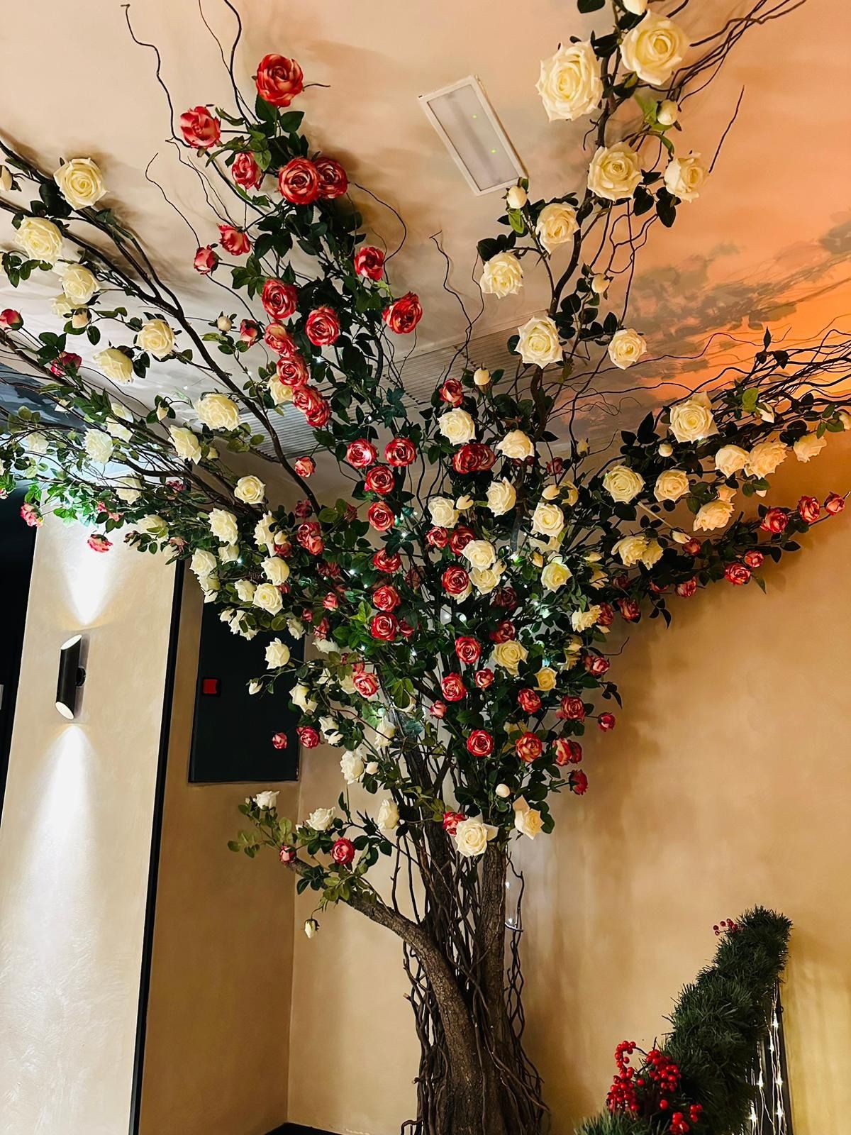 Árbol artificial con rosas rojas y color crema, que se extiende desde el suelo hasta el techo en una habitación bien iluminada.