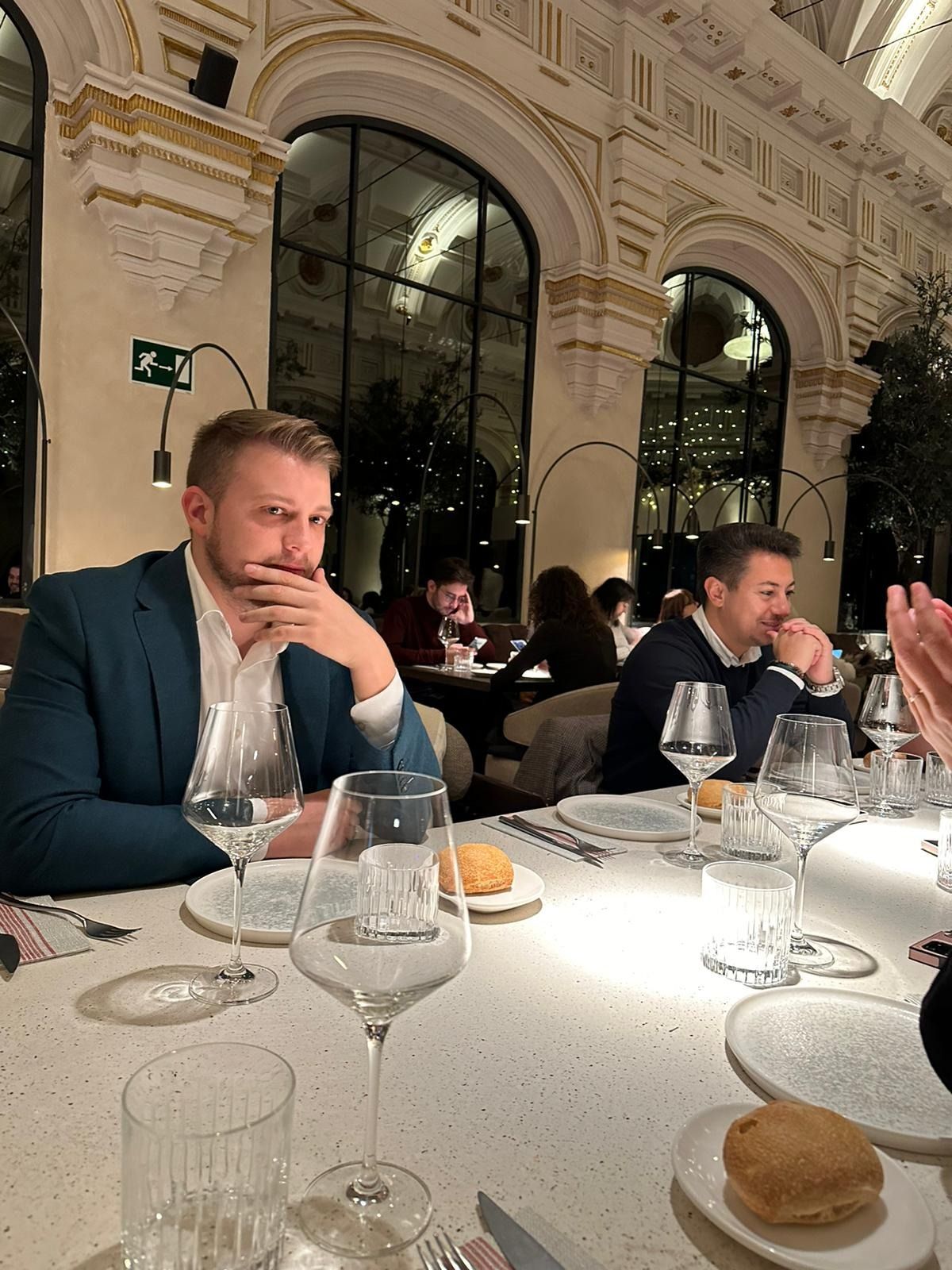 Dos hombres sentados a la mesa de un restaurante elegante; uno de ellos lleva una chaqueta azul verdosa que le cubre la boca. Ambiente elegante con grandes ventanales.