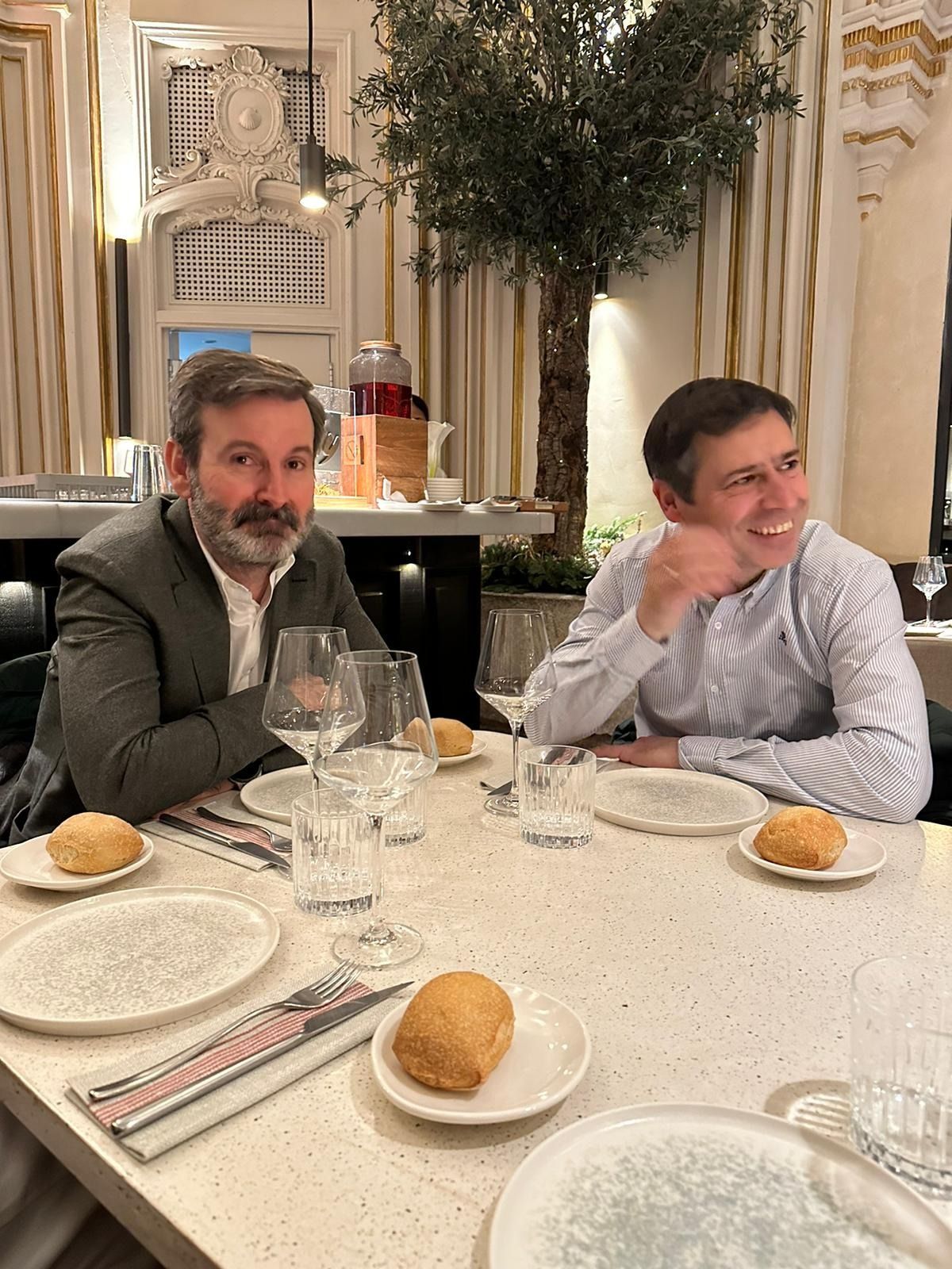 Dos hombres sentados a la mesa de un restaurante. Uno sonríe, el otro mira a la cámara. Hay panecillos y vasos sobre la mesa.