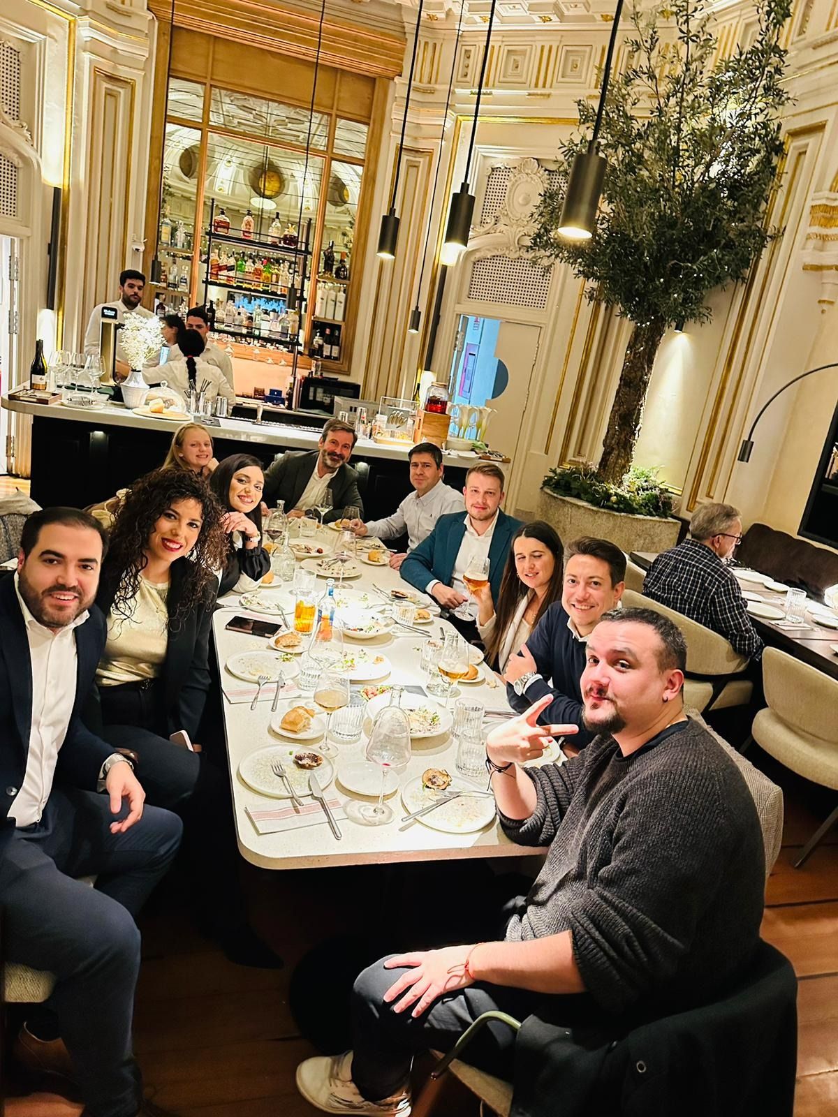 Grupo de personas sentadas a una mesa larga en un restaurante ornamentado; algunas sonríen, otras señalan.