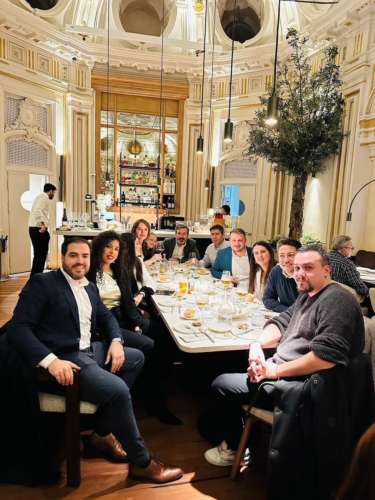 Grupo de personas sentadas a una mesa larga en un comedor ornamentado, algunas sonriendo.