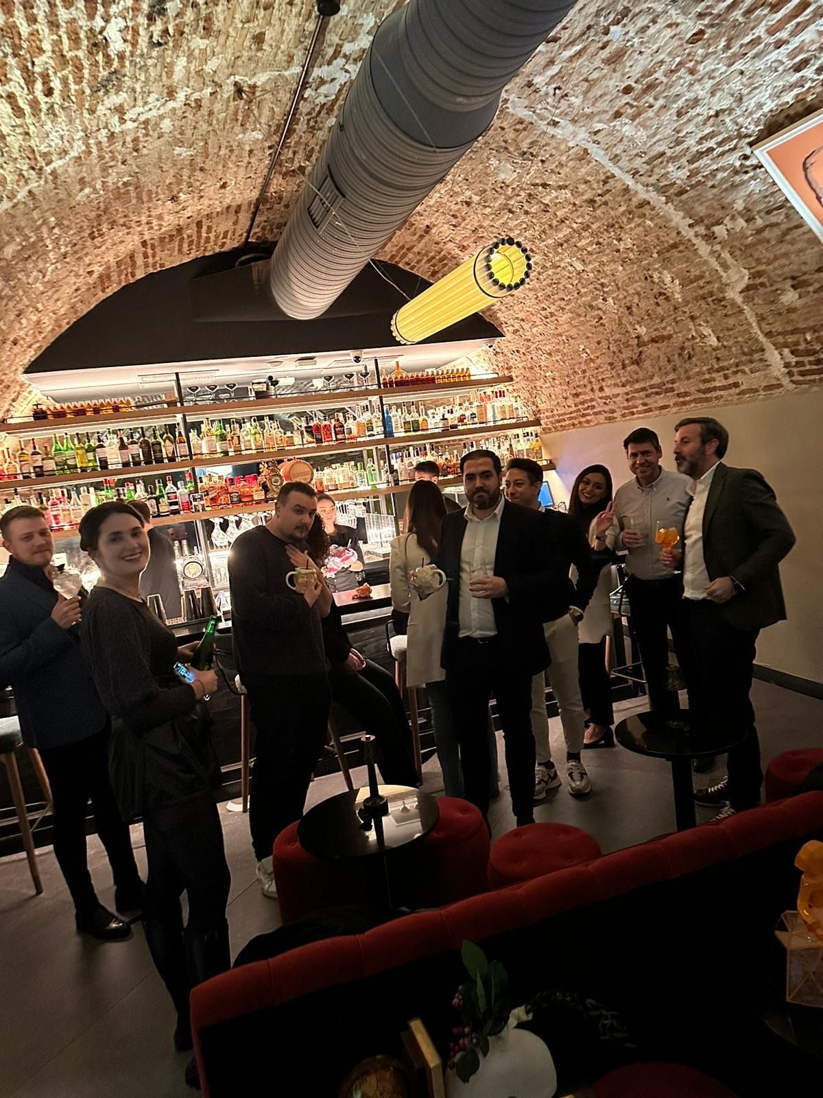 Grupo de personas en un bar tomando algo, bajo un techo abovedado de ladrillo. Sonríen y charlan.