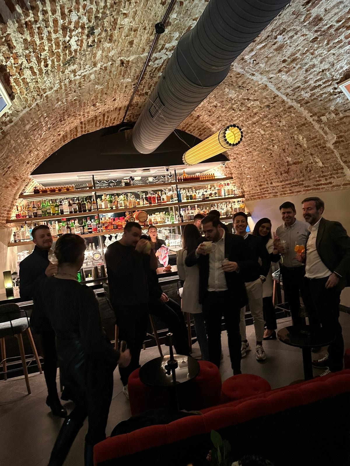 Gente en un bar con techo abovedado de ladrillo. Botellas alineadas al fondo. Algunos llevan bebidas.