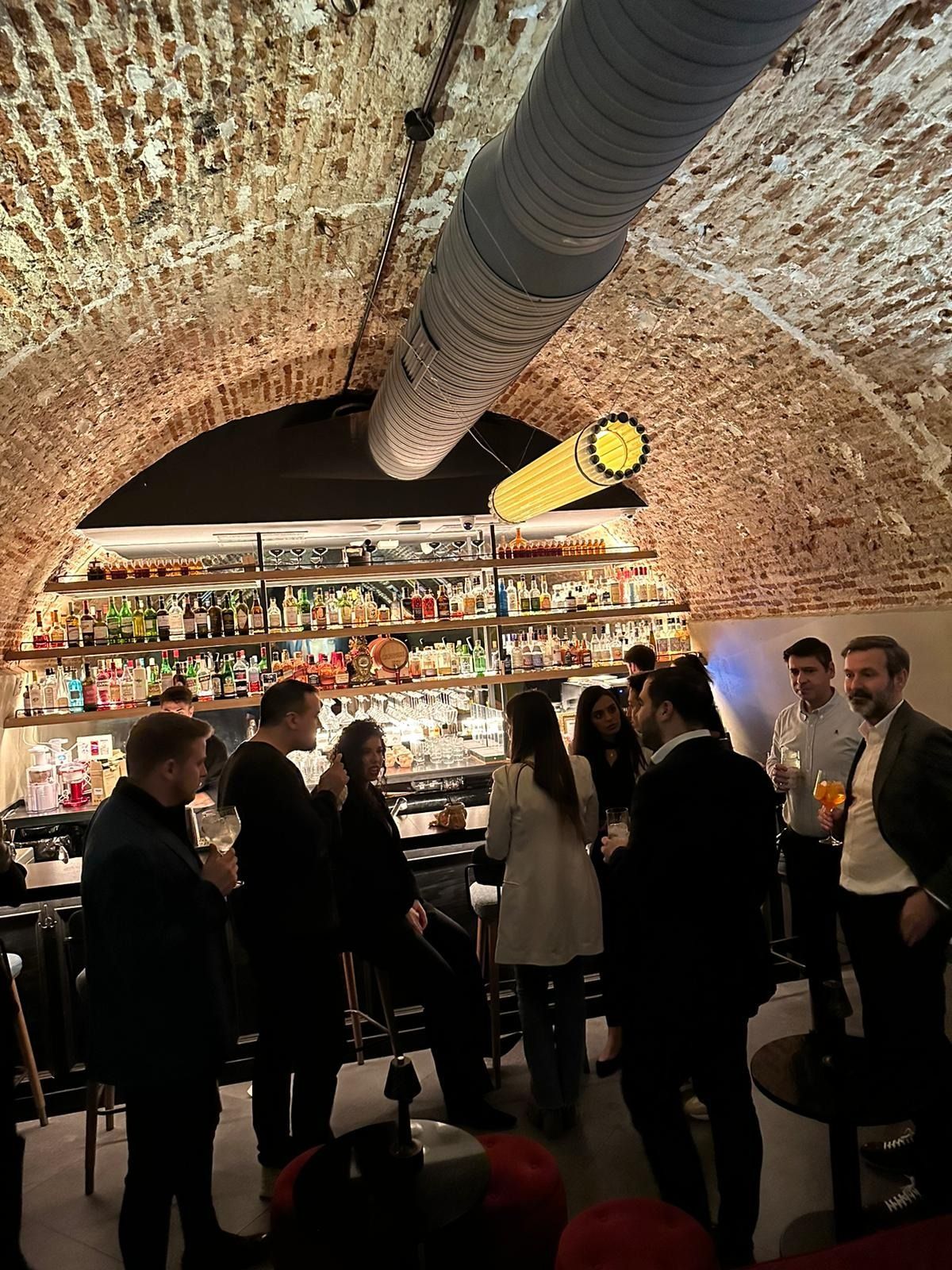 Gente en un bar con techo abovedado de ladrillo. Botellas alineadas en la pared del fondo, y un conducto de estilo industrial la recorre.