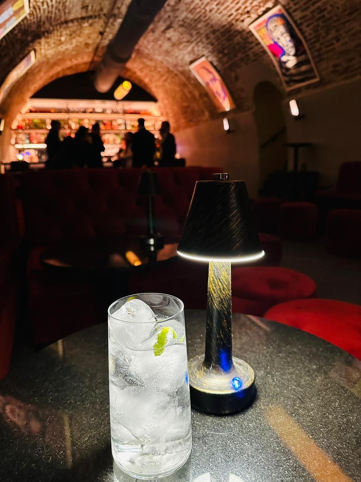 Un cóctel y una lámpara sobre una mesa en un bar con poca luz; al fondo, las siluetas de los clientes.