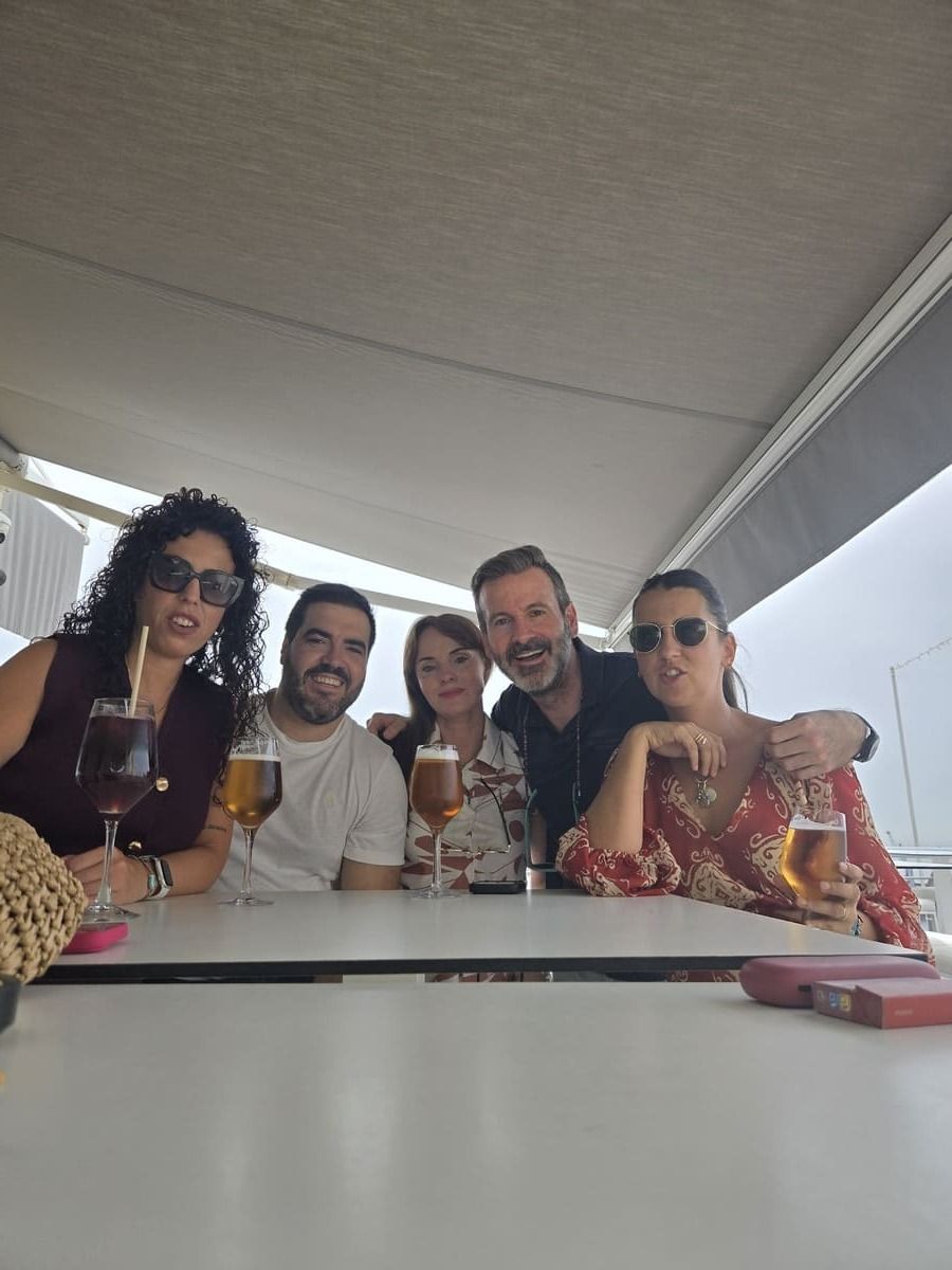 Cinco personas sentadas a una mesa, bebiendo y sonriendo. Ambiente al aire libre, bajo una pérgola.
