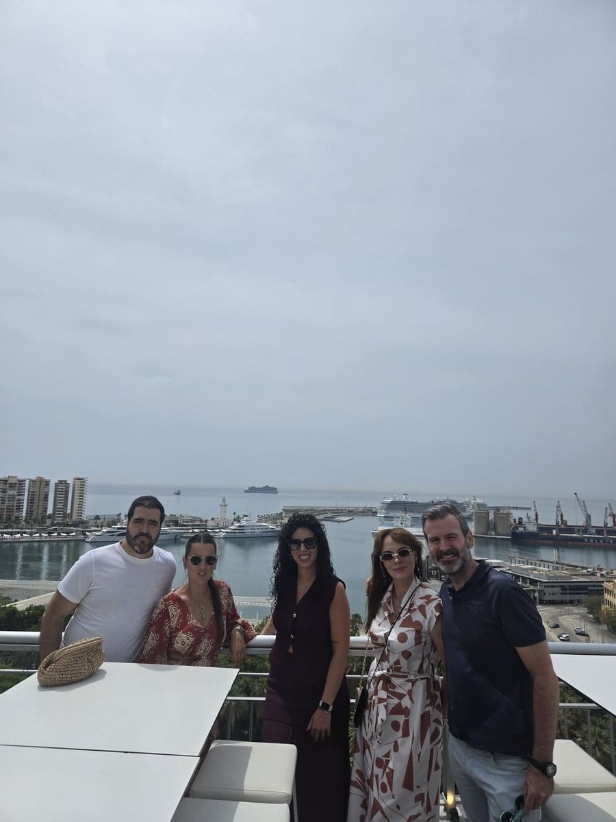 Cinco personas sonriendo en una azotea con vistas a un puerto, cielo nublado.