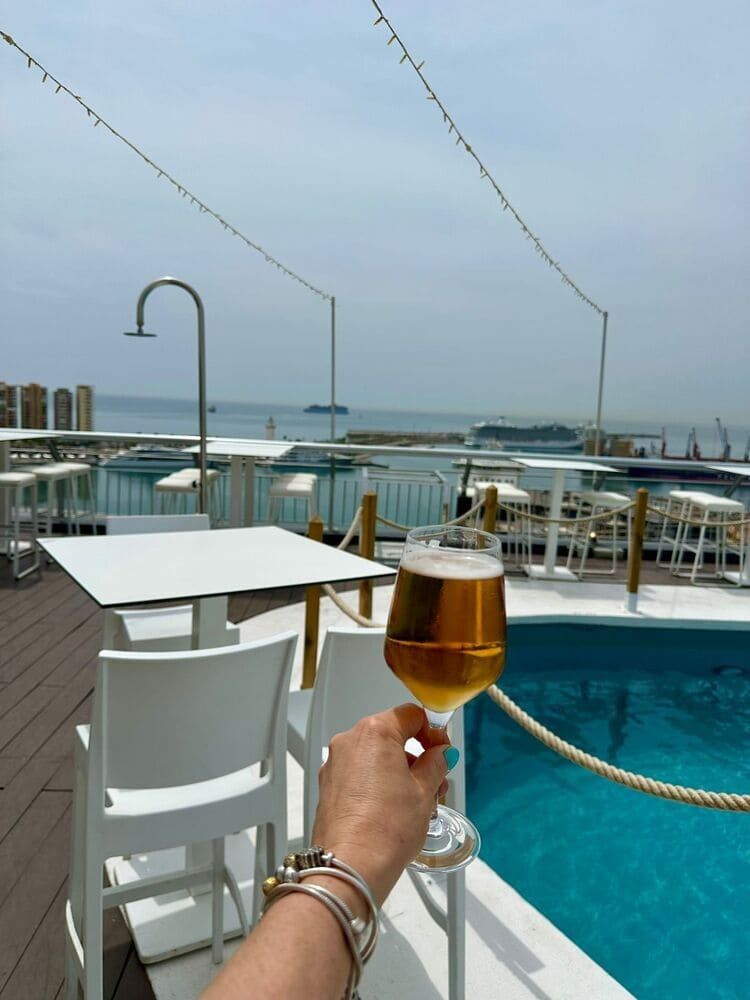 Mano sosteniendo un vaso de cerveza, con vistas a una piscina y al mar, en un entorno soleado al aire libre.