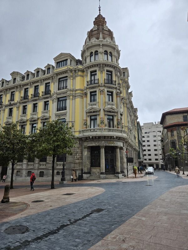 Edificio esquinero amarillo ornamentado con cúpula, árboles y calle en un día nublado.
