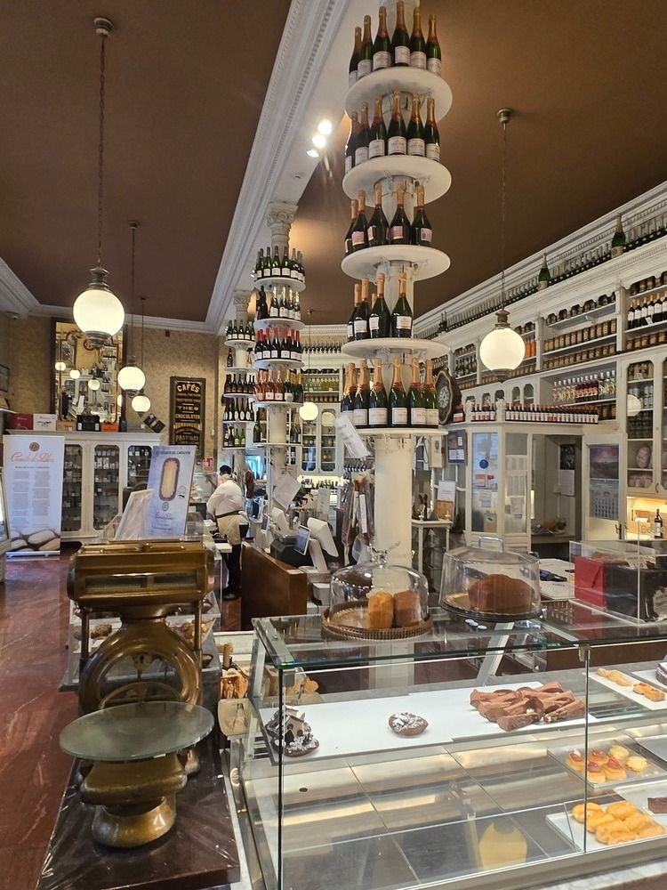 Interior de una tienda con botellas de vino expuestas en estantes y en torres colgantes; productos horneados en una vitrina.