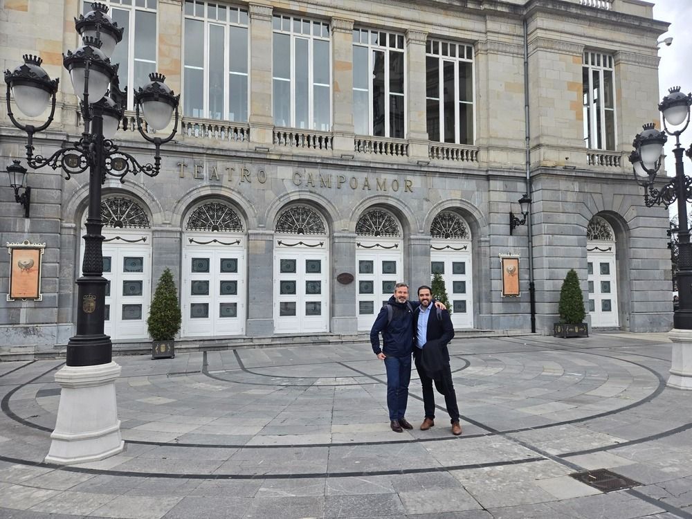 Dos hombres están de pie frente al edificio del Teatro Campoamor, bajo farolas ornamentadas. Edificio de piedra gris, puertas blancas.