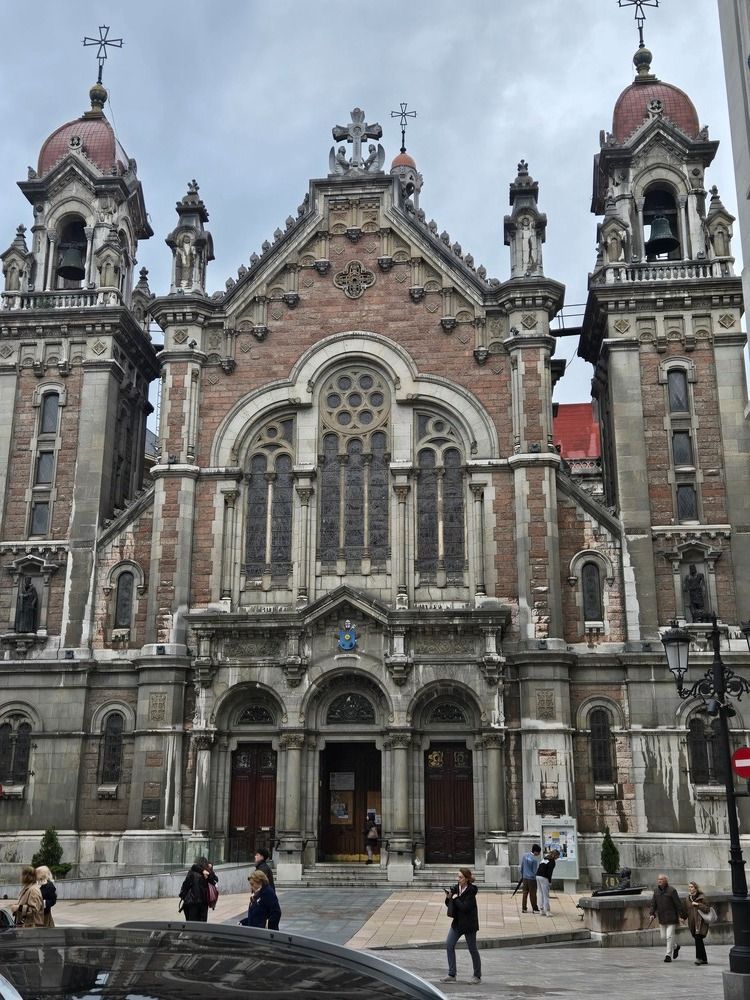 Gran iglesia ornamentada con dos campanarios. Gente caminando cerca de la entrada.