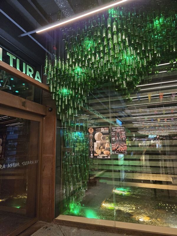Entrada a un local comercial con fachada de cristal, adornada con botellas de cristal verde colgantes.