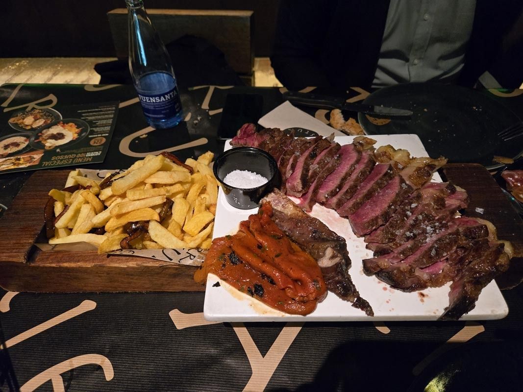 Cena de bistec: bistec en rodajas, papas fritas, pimientos y salsa en una mesa con una botella de agua y menú.