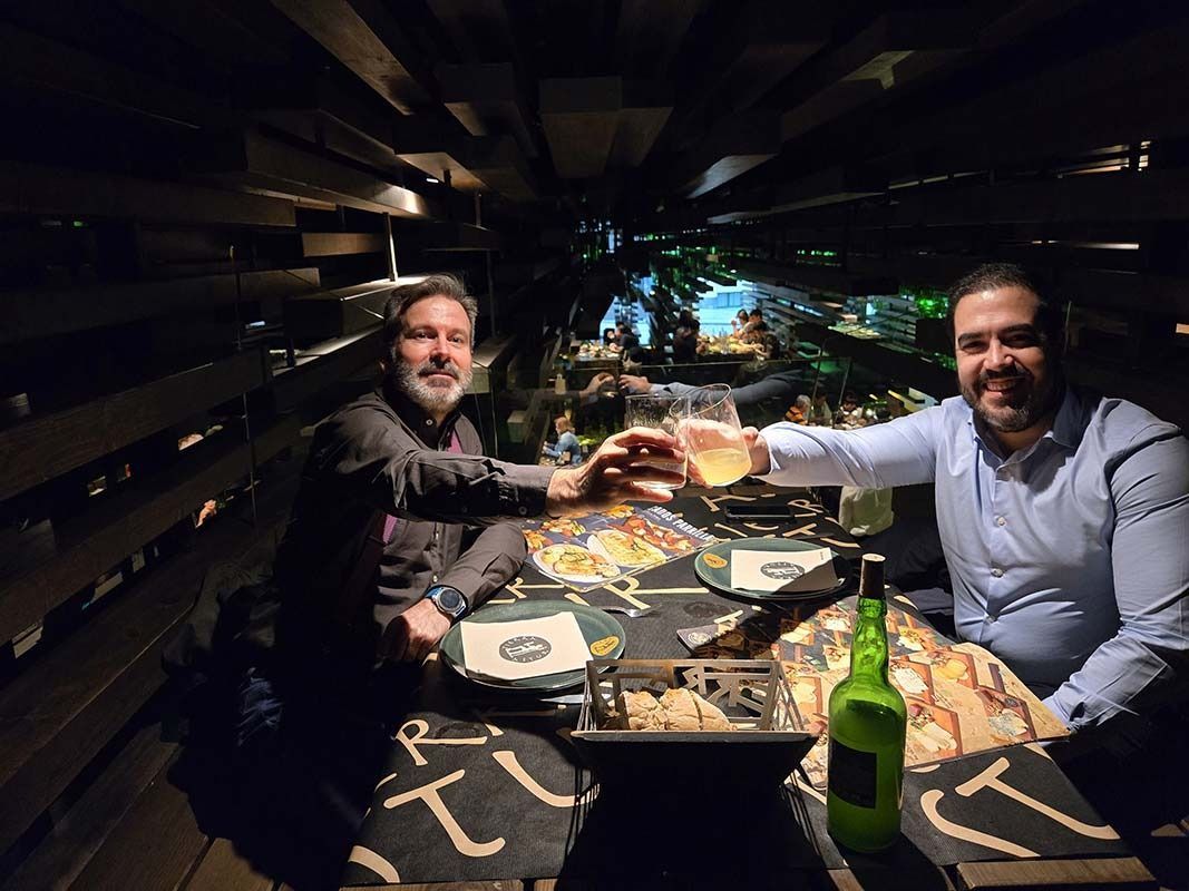 Dos hombres brindando con unas copas en una mesa con paneles de madera en un restaurante con poca luz.
