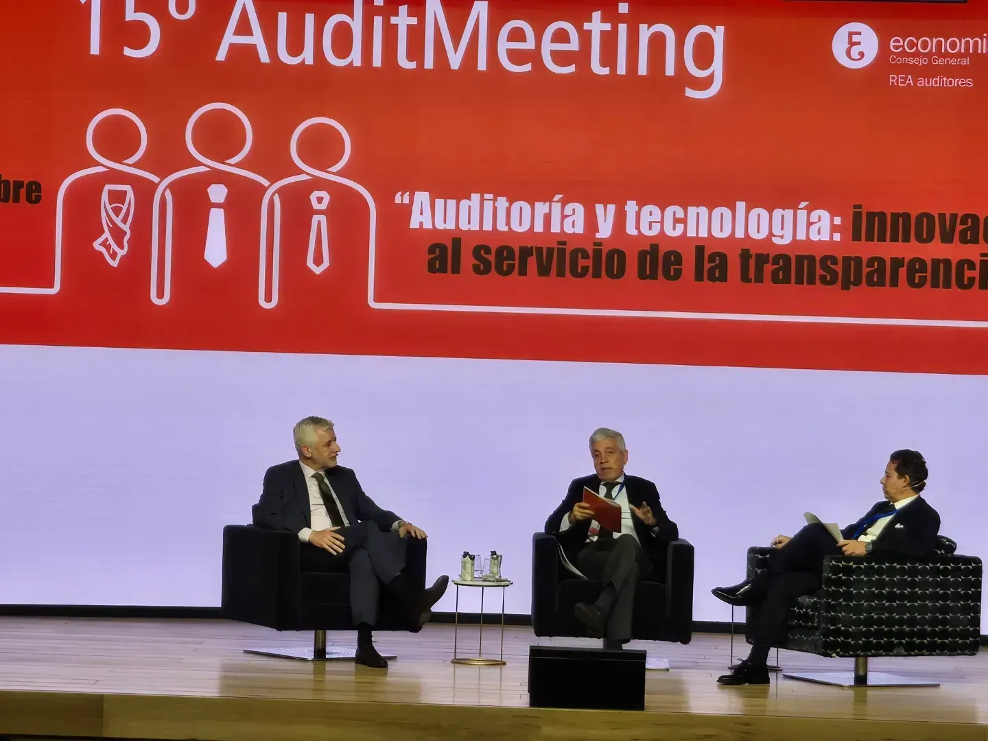 Tres hombres sentados en el escenario durante una conferencia, hablando. Pancarta roja con texto.