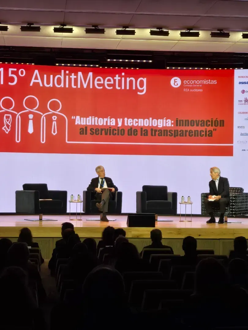 Público en la conferencia; dos hombres sentados en el escenario bajo una pancarta que dice 
