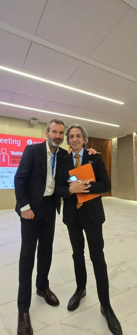 Dos hombres con traje posan juntos en el interior, uno de ellos con una carpeta naranja.