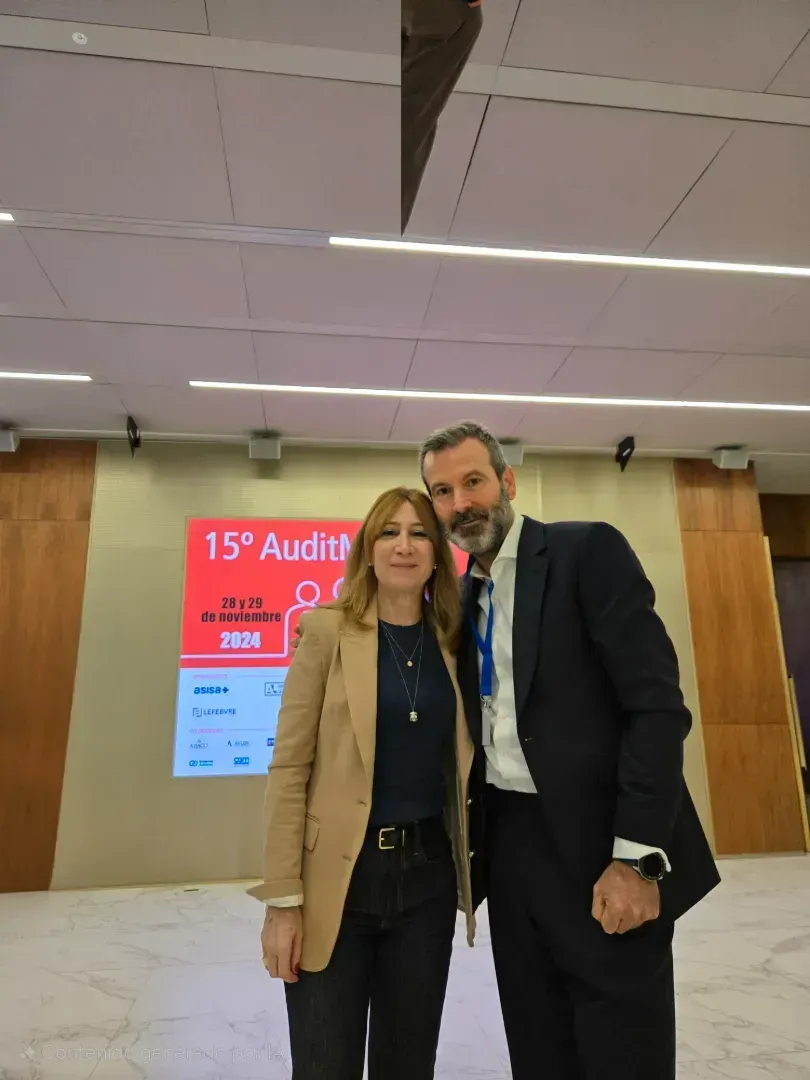 Dos personas de pie frente a una pantalla. Mujer con chaqueta color beige, hombre con traje. Ambiente interior con un letrero.