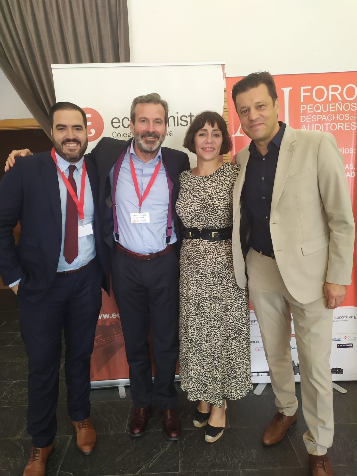 Cuatro personas posando juntas en un evento. Están de pie cerca, sonriendo, con vestimenta formal, frente a una pancarta.