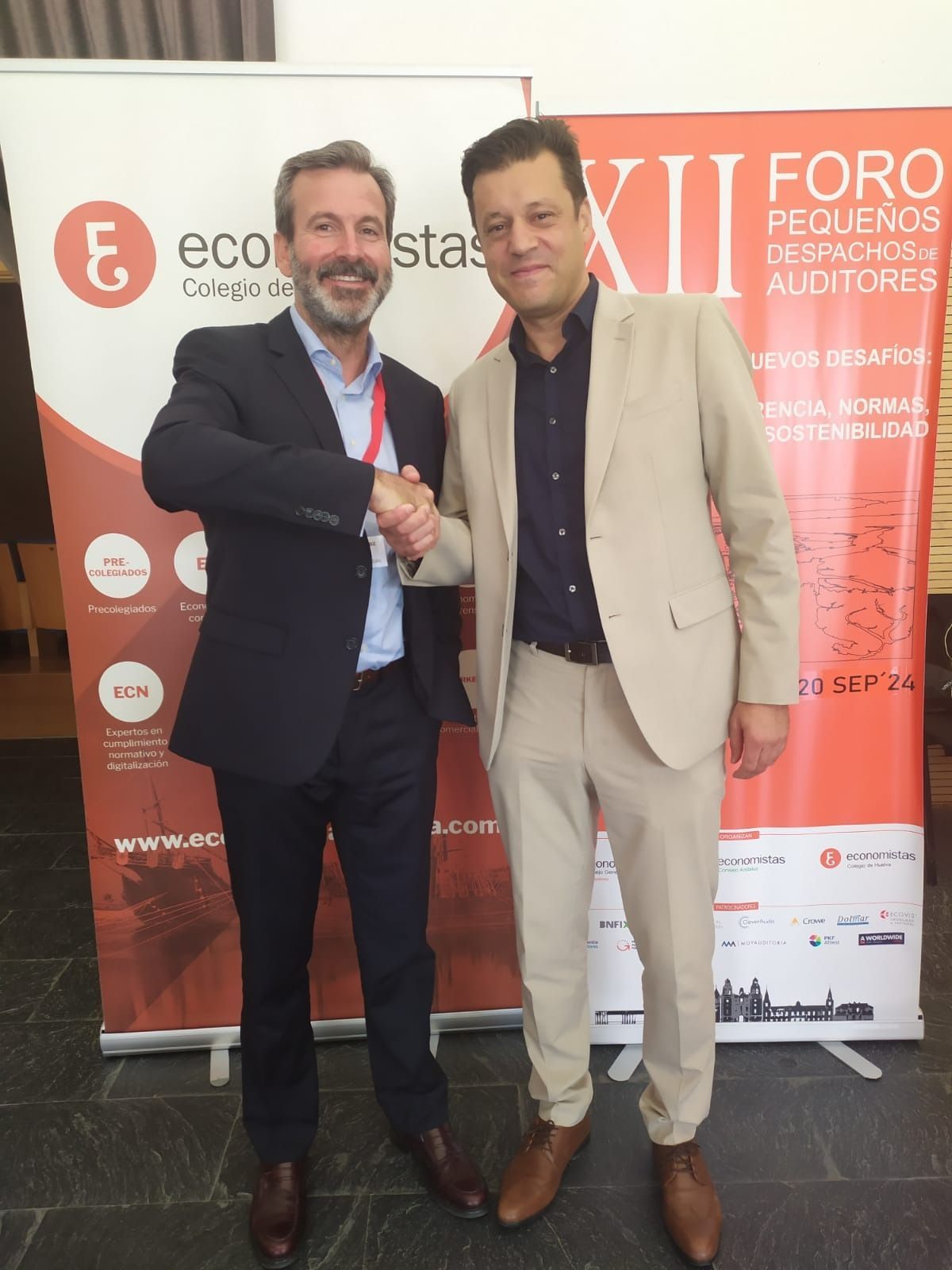 Dos hombres se dan la mano frente a una pancarta en un foro. Uno lleva traje, el otro una chaqueta clara.