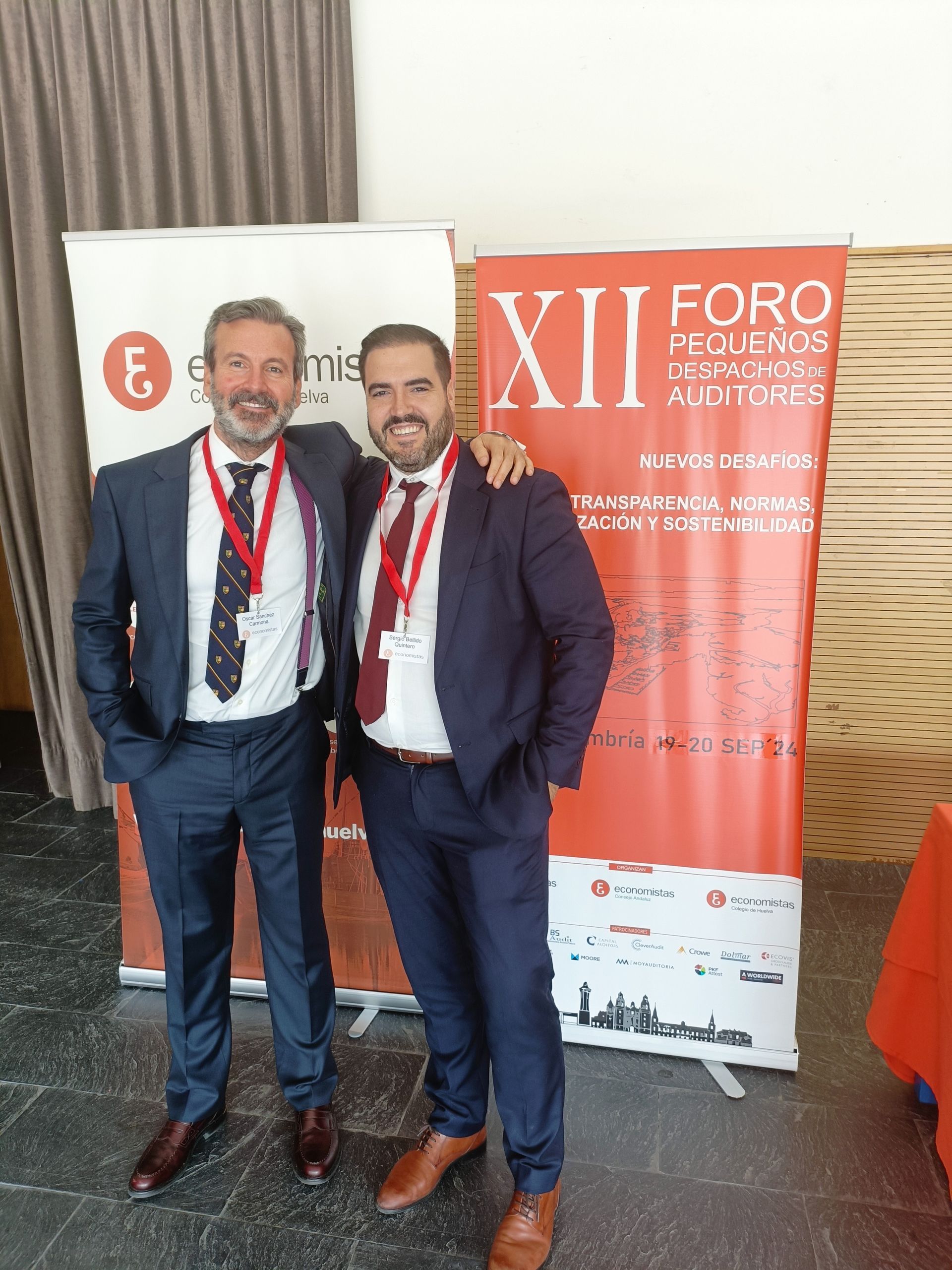 Dos hombres de traje posan en una conferencia. Están de pie frente a las pancartas del evento, sonriendo.