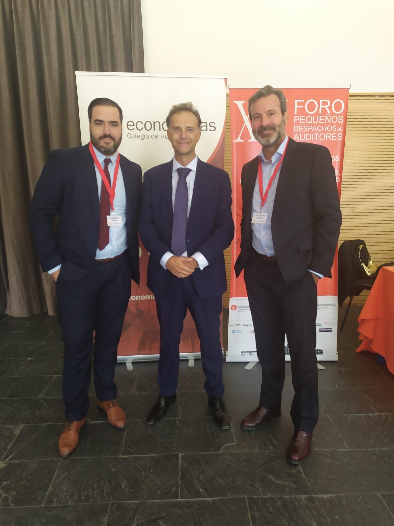 Tres hombres de traje posan delante de pancartas. Dos llevan cordones rojos.