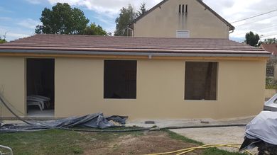 Maison de plain-pied beige avec toit marron et fenêtres condamnées, vue de face dans une cour.