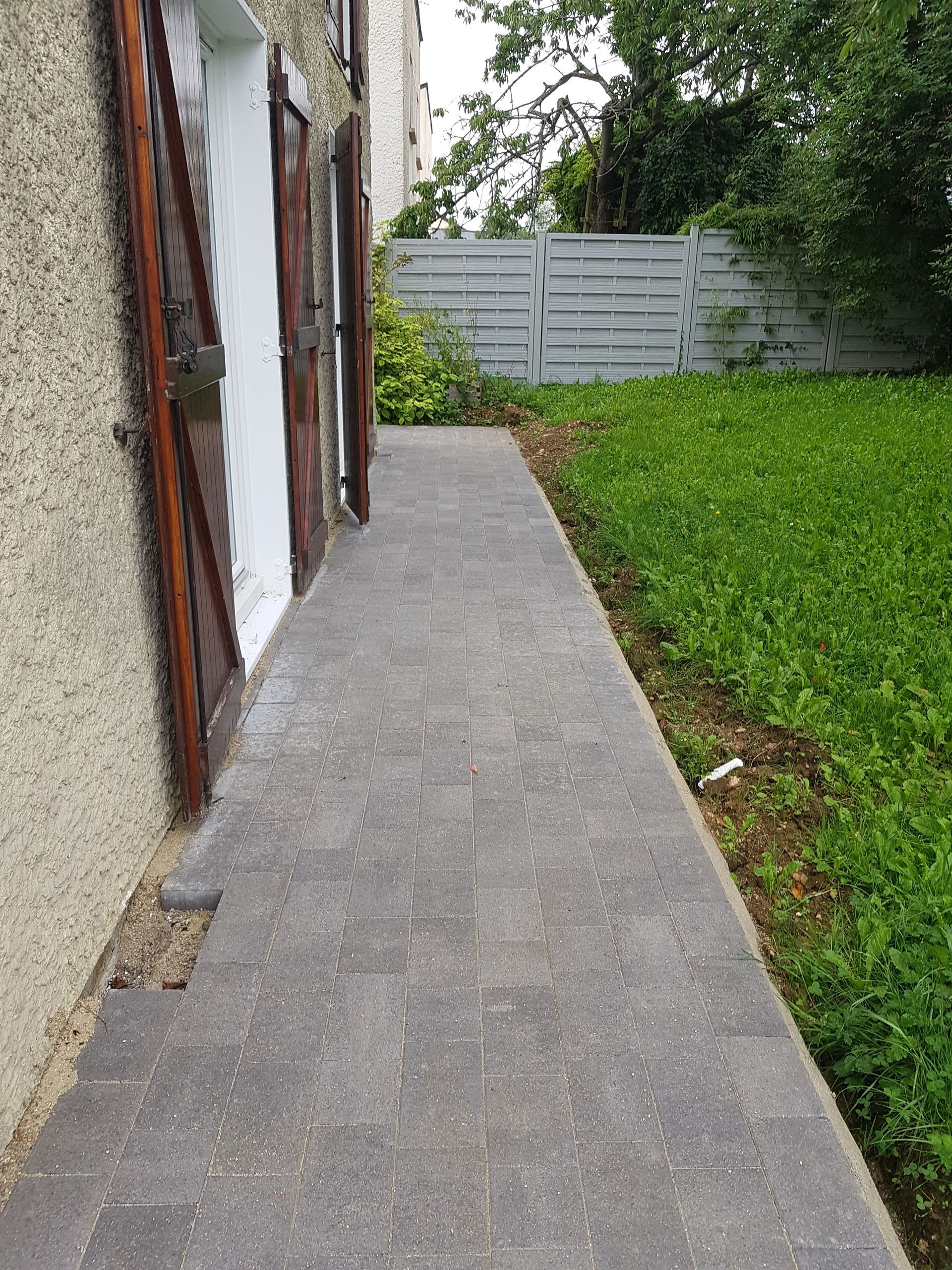 Étroite allée en béton longeant une maison et menant à une clôture grise et une pelouse verte.
