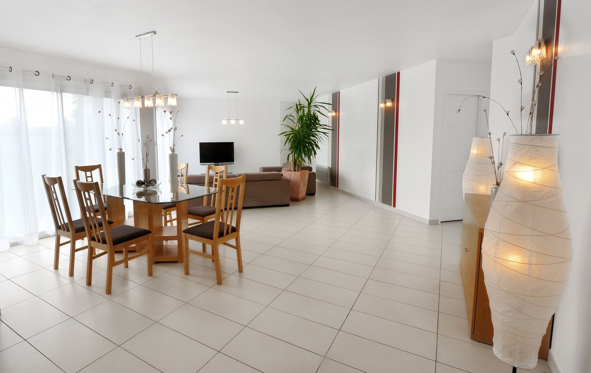 Salle à manger lumineuse et moderne avec table et chaises en bois, sol carrelé blanc et lampadaires hauts.