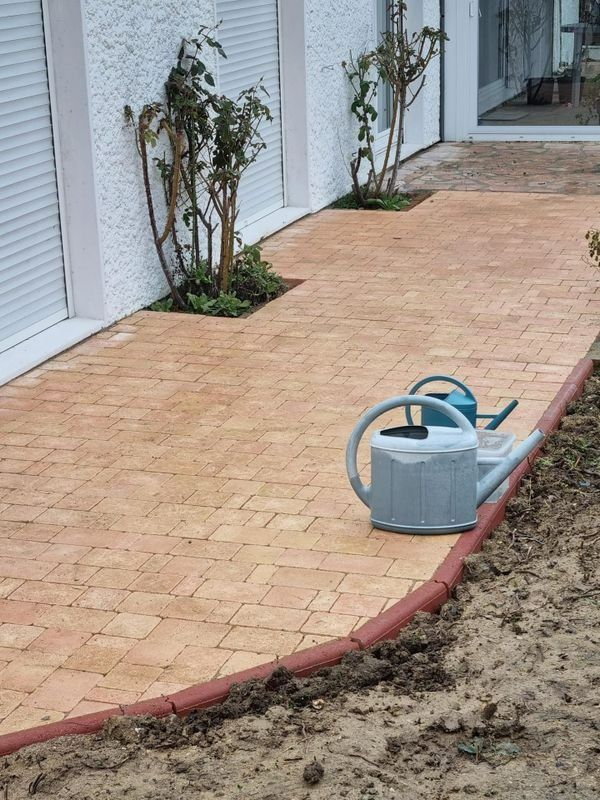 Arrosoir sur une terrasse en briques à côté d'un bâtiment blanc et de petites plantes, avec de la terre nue au premier plan.