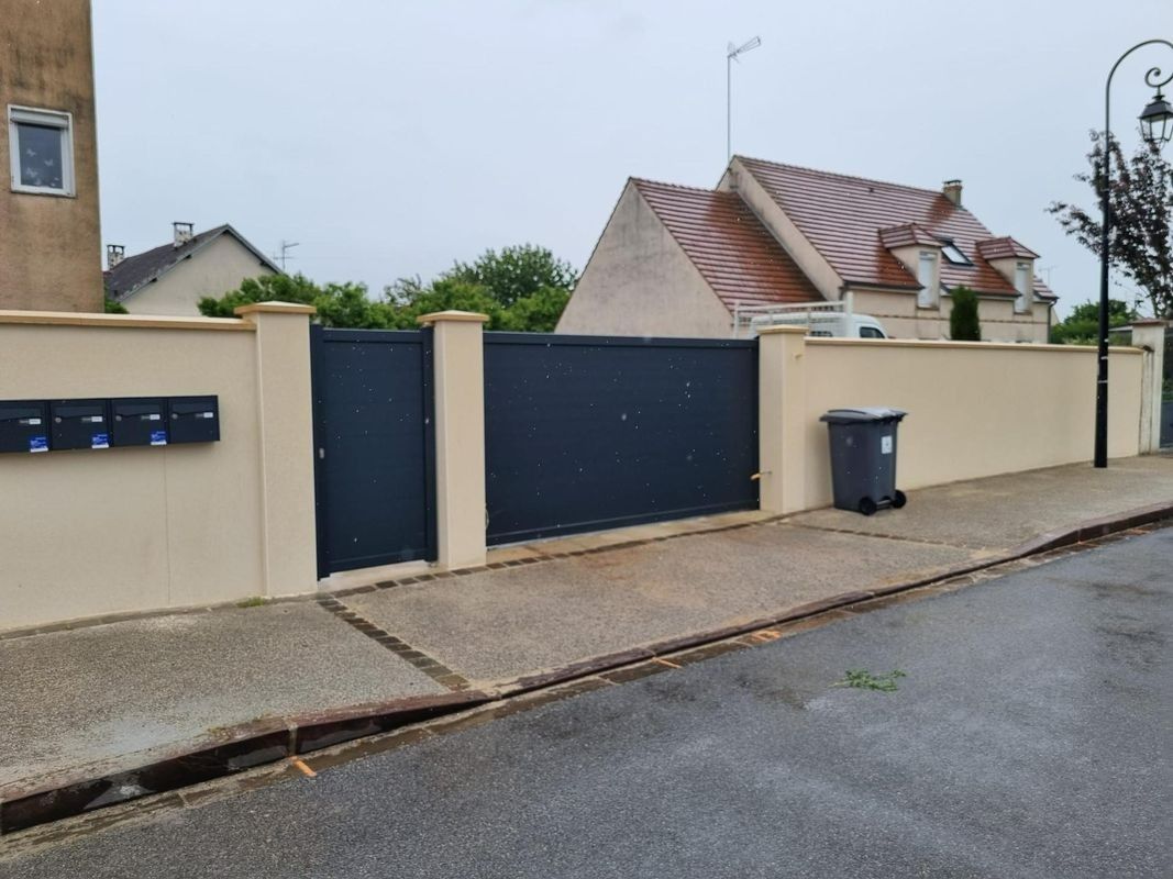 Entrée d'une maison individuelle aux murs beiges, portail sombre, boîte aux lettres et poubelle donnant sur une rue mouillée.
