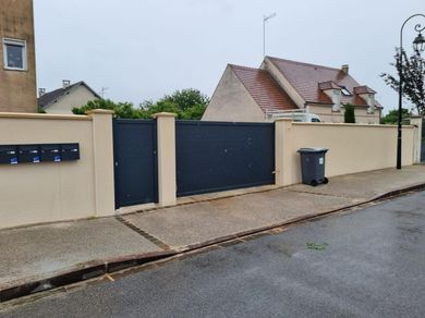 Entrée d'une maison individuelle aux murs beiges, portail sombre, boîte aux lettres et poubelle donnant sur une rue mouillée.