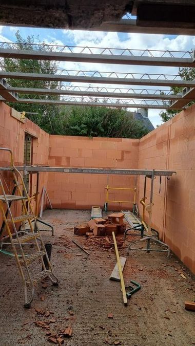 Pièce en briques partiellement démolie, avec poutres de toit apparentes, échelles et débris de construction éparpillés.