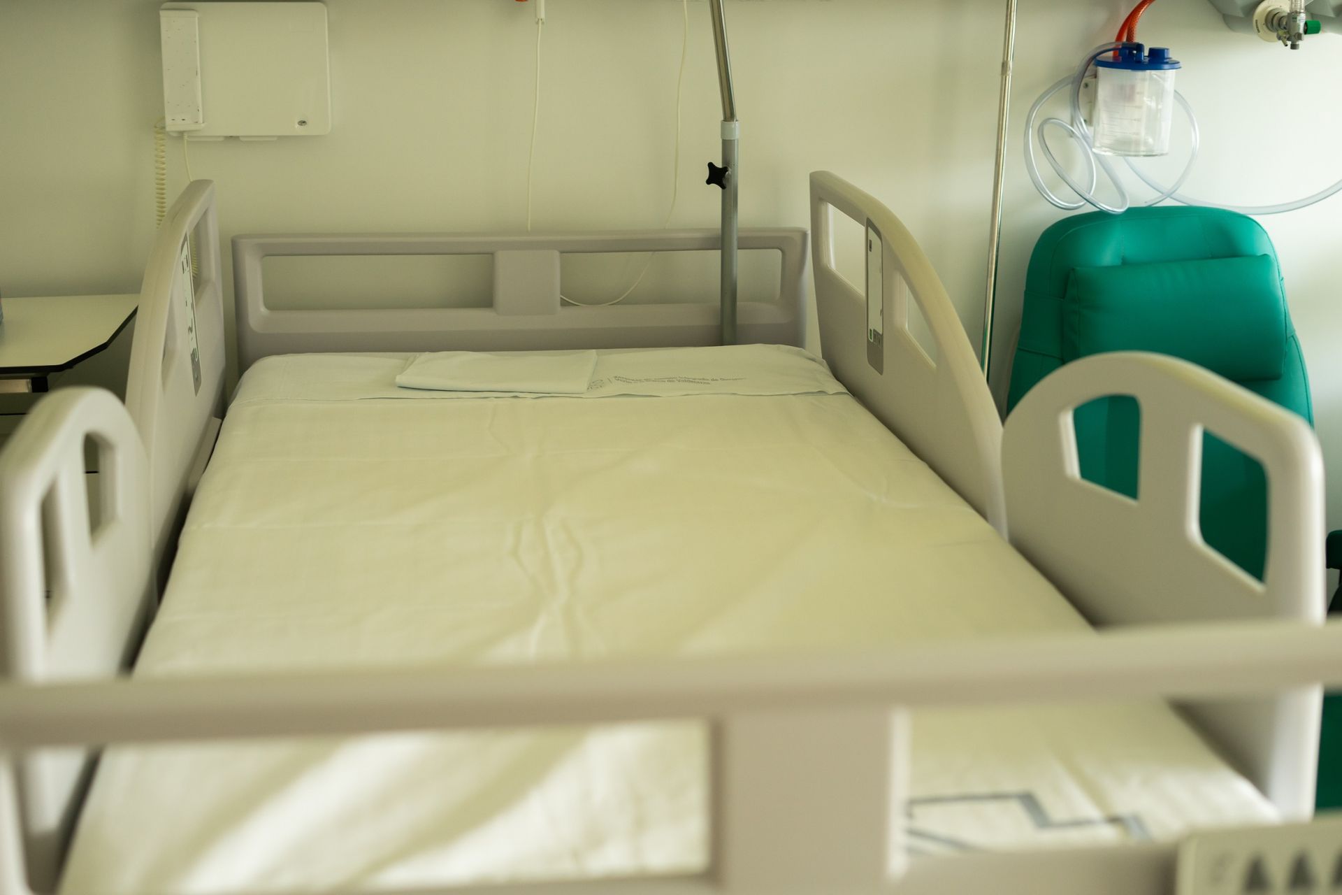 Una cama de hospital vacía con una silla verde al fondo.