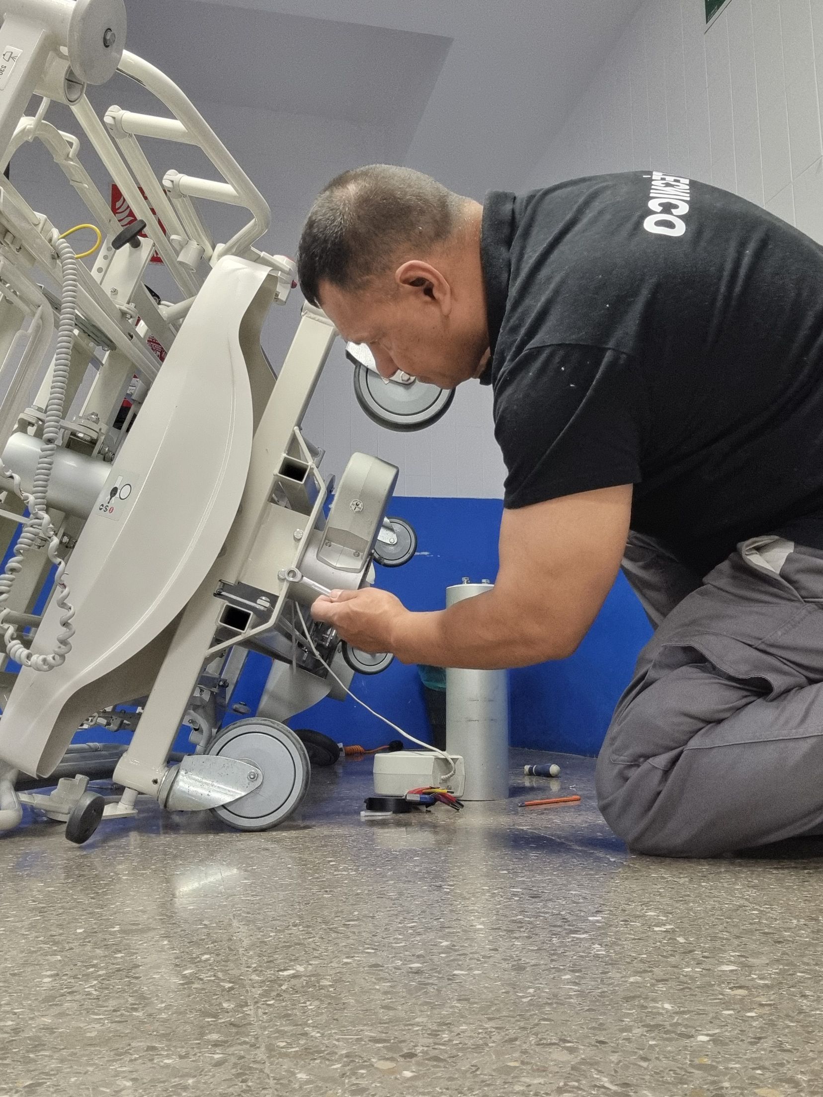 Técnico arrodillado junto a una máquina médica, ajustando un componente en una sala clínica.