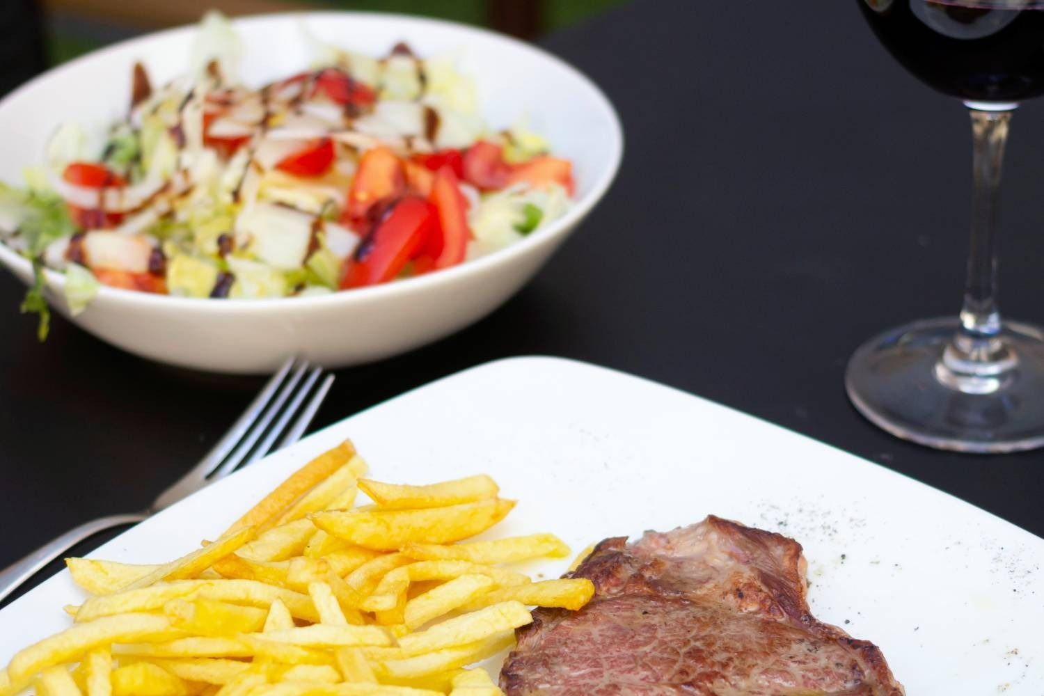 Un plato de carne y patatas fritas junto a un bol de ensalada y una copa de vino.
