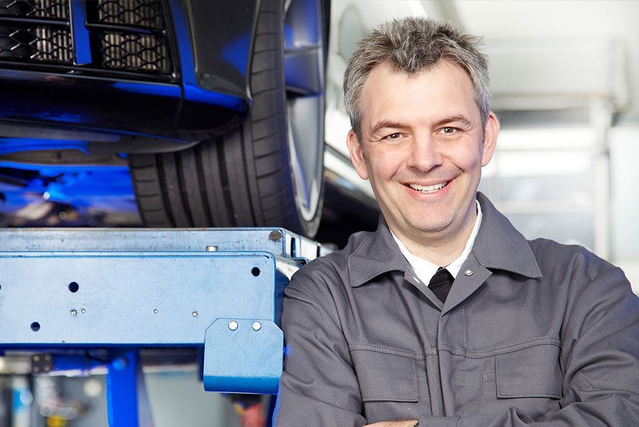 Ein lächelnder Mechaniker steht vor einem Auto auf einer Hebebühne.