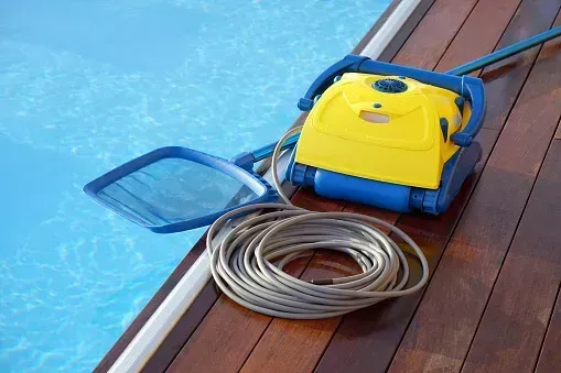 Robot amarillo limpiafondos con manguera en una terraza de madera junto a agua azul.